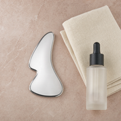 Stainless-steel gua sha beside face oil and an ivory towel