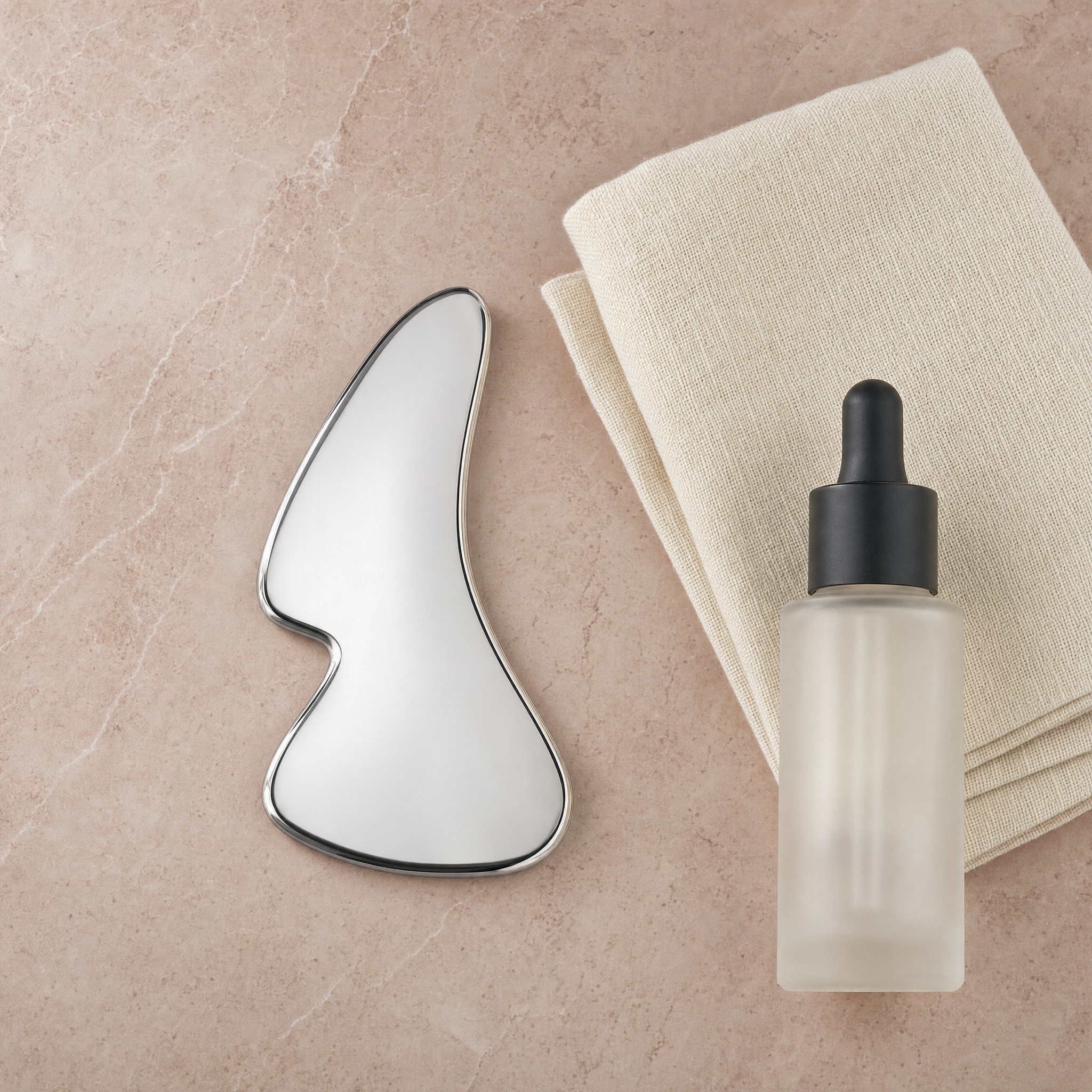 Stainless-steel gua sha beside face oil and an ivory towel